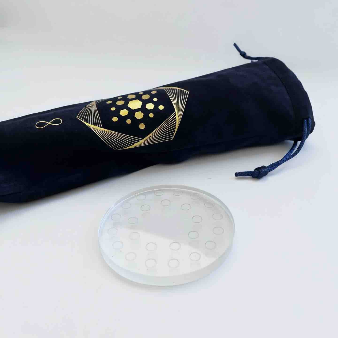 A round clear crystal clearing stand with small indents sits beside a navy blue drawstring pouch with gold geometric designs, both from The Wellness Enterprise's Aqua Energizer Care Kit.