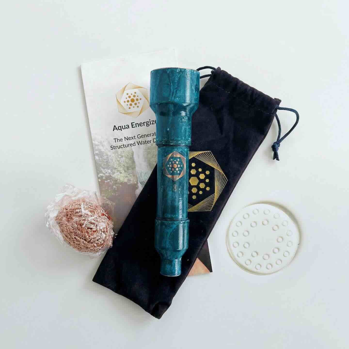 The Aqua Energizer On-the-Go Structured Water Device by Aqua Energizer includes a blue cylinder, black drawstring pouch, perforated white disc, mesh-wrapped ball, and leaflet—all arranged on a white surface for enhanced cellular hydration.