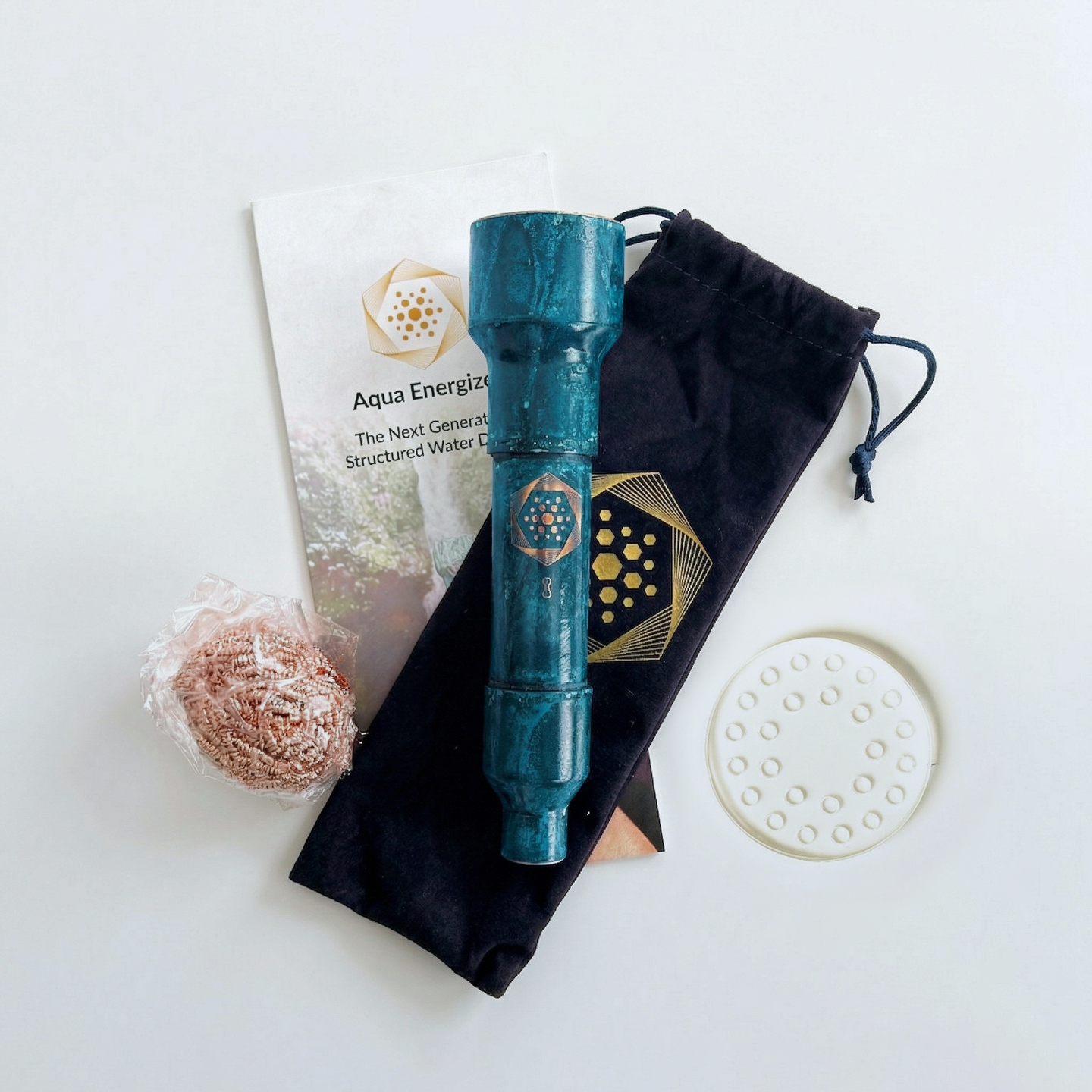 The Aqua Energizer Care Kit by The Wellness Enterprise includes a blue cylindrical device with a gold hexagon logo, black drawstring pouch, copper scrubber, crystal clearing stand, white disk, and care kit brochure arranged on a white surface.