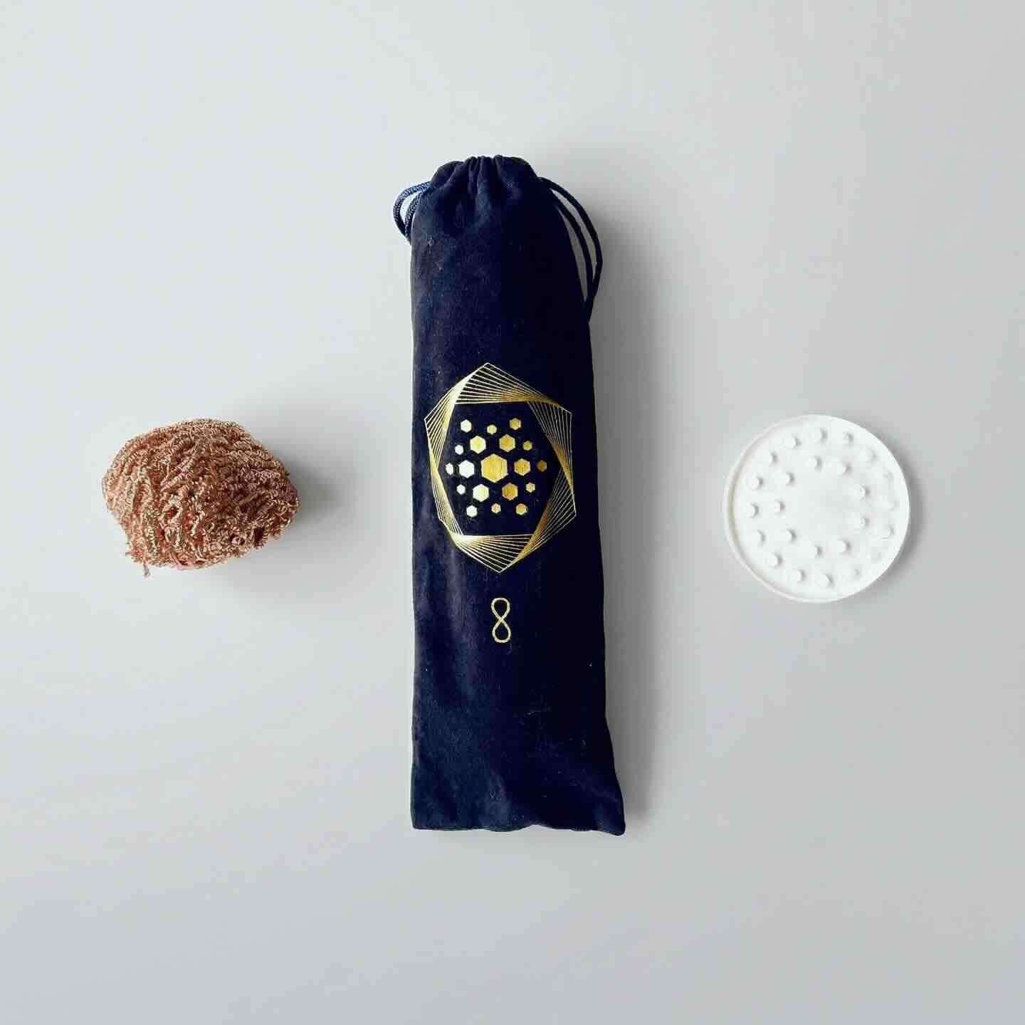 The Aqua Energizer Care Kit by The Wellness Enterprise features a brown textured item, a black drawstring pouch with a gold geometric pattern, and a crystal clearing stand arranged side by side on a light surface.