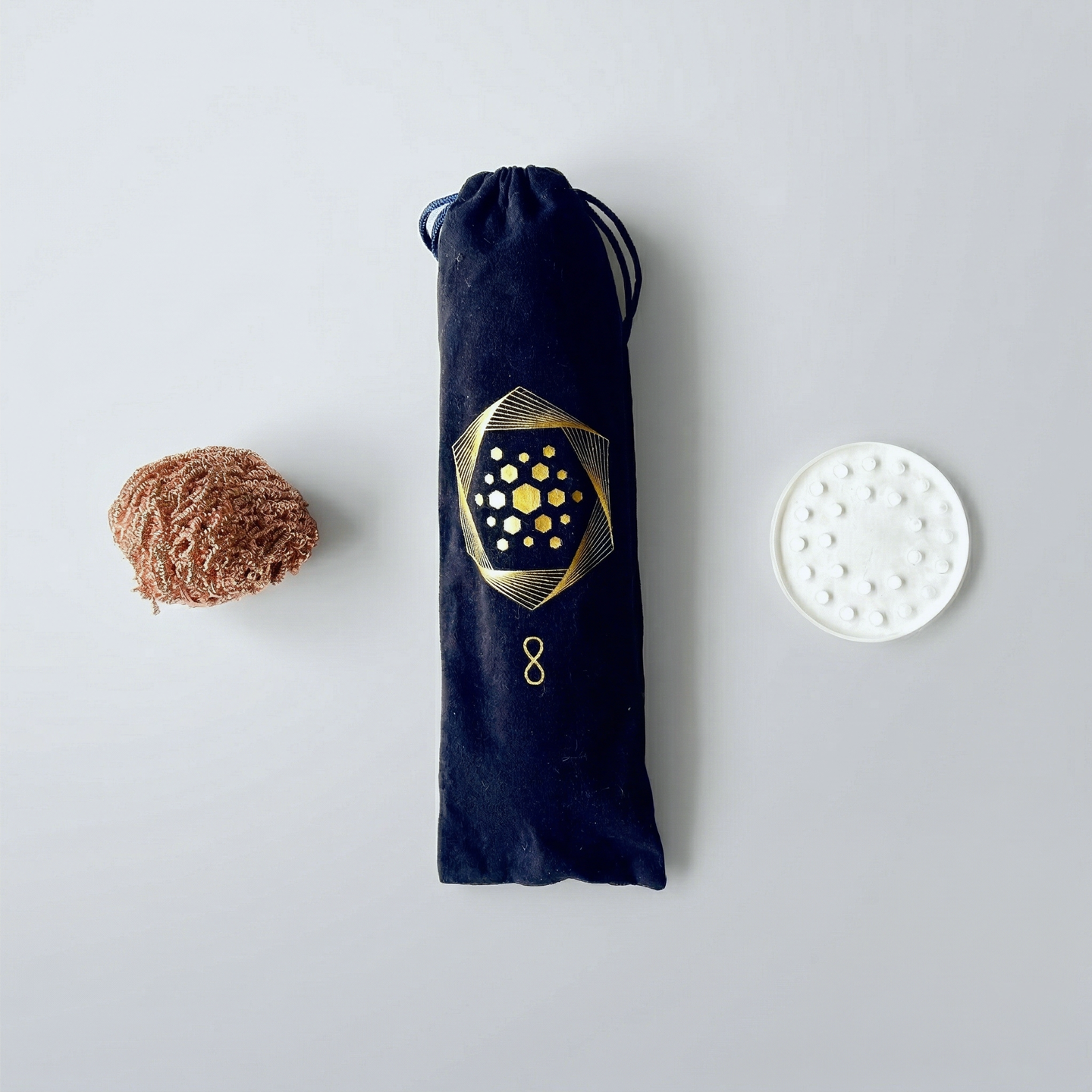 The Wellness Enterprise’s Aqua Energizer Care Kit features a brown sponge, a black drawstring pouch with gold geometric design, and a white round ceramic dish with raised dots, arranged on a light background.