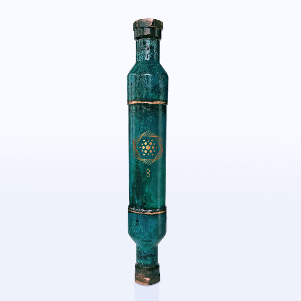 A metallic, cylindrical green device with brass-colored edges and a gold geometric symbol in the center—identified as The Wellness Enterprise’s Whole House Aqua Energizer + FREE Aqua Energizer On-the-Go structured water device—stands upright on a white background, designed to support cellular hydration.
